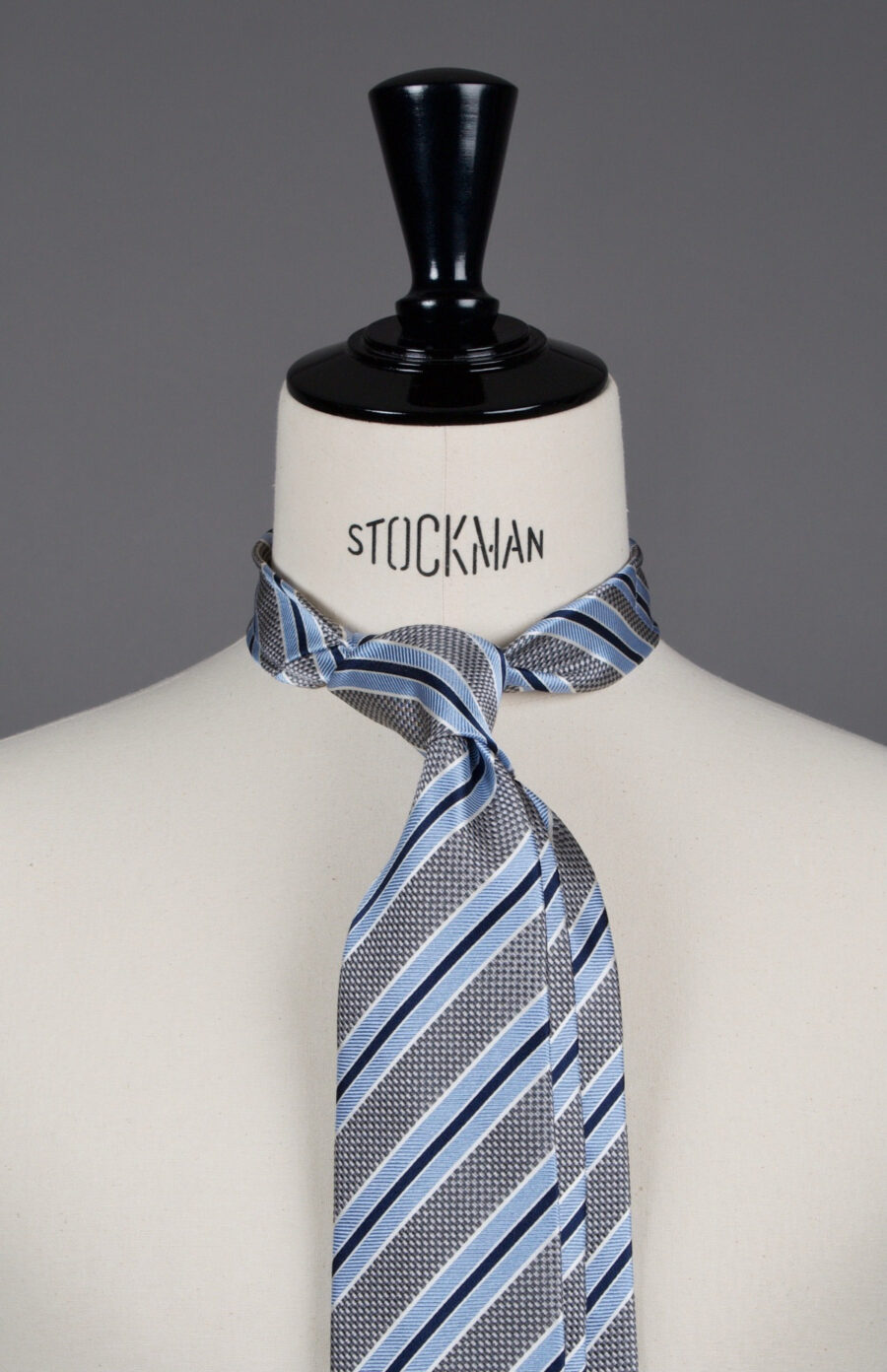 Cravate sartoriale à 3 plis, en soie tissée Jacquard, grise rayée bleu et aperçu du noeud simple sur buste Stockman.