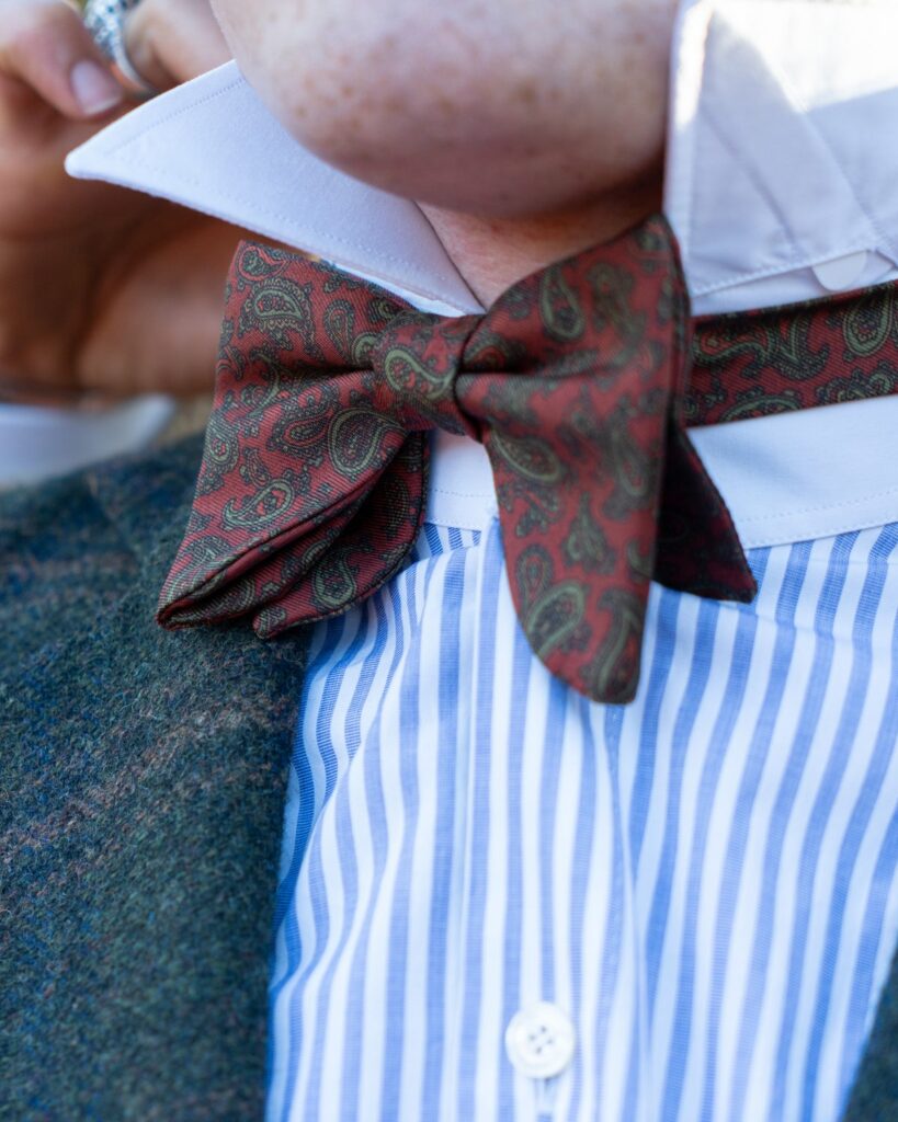 Nœud papillon en soie à motif cachemire bordeaux et vert, sur une chemise rayée bleue et une veste en tweed